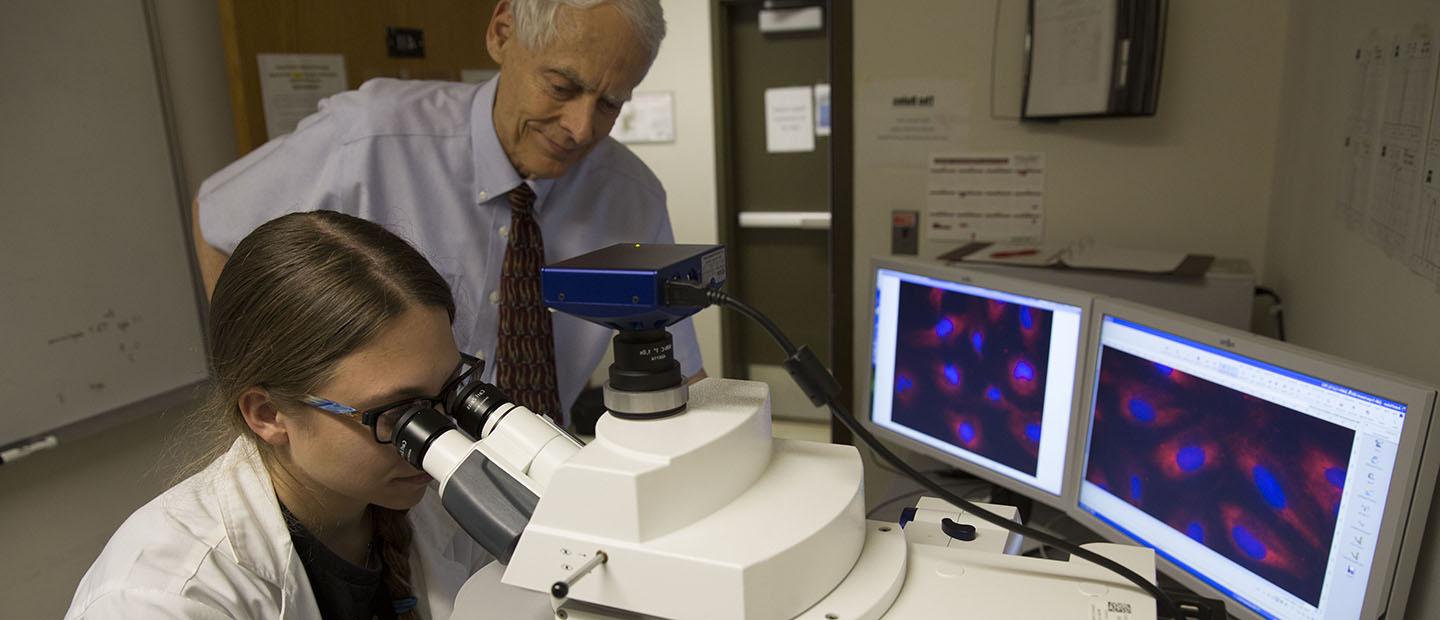 professor standing over a student looking in a microscope with two computer