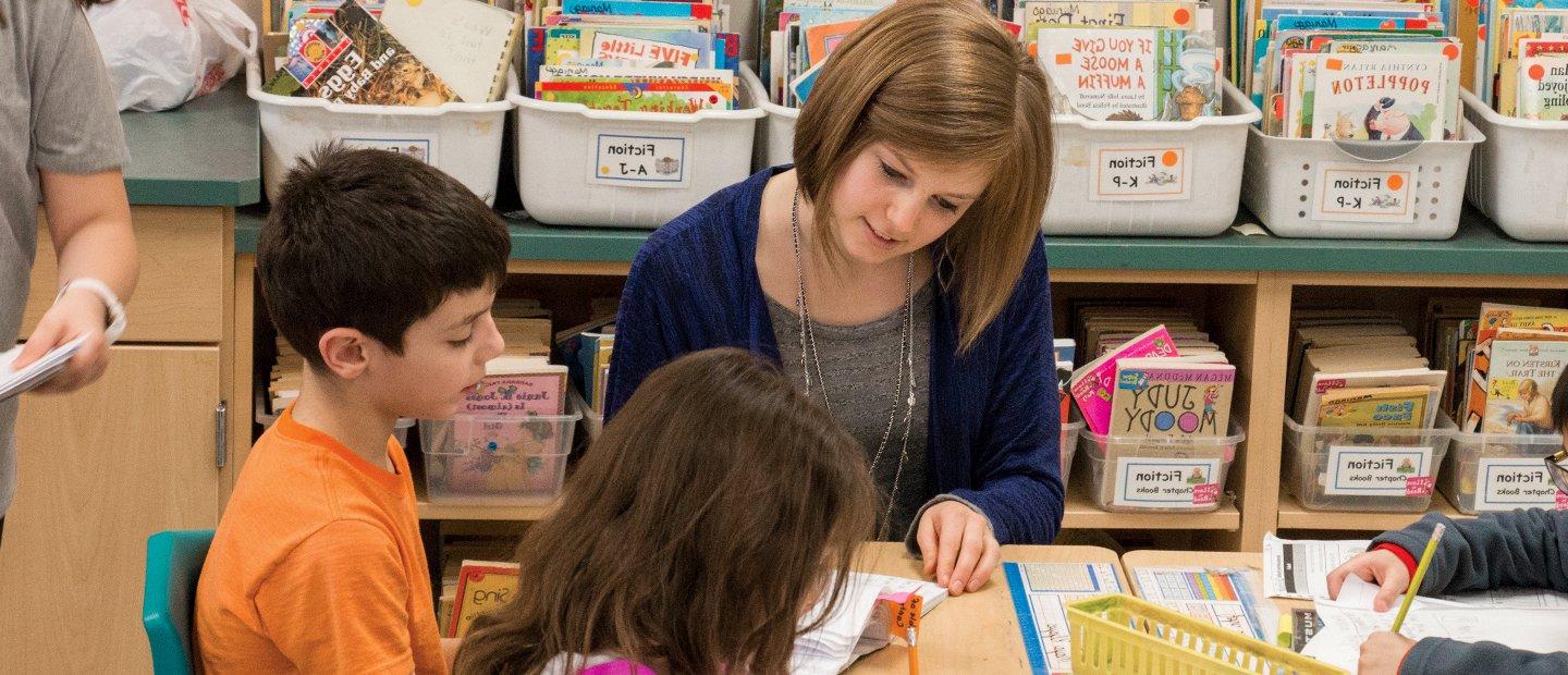 A female teacher seated with young students, reading books.